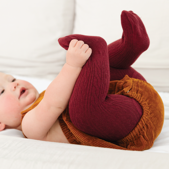 Baby lying down wearing Little Stocking Co. Burgundy Cable Knit Tights, cozy cable knit tights for baby girl