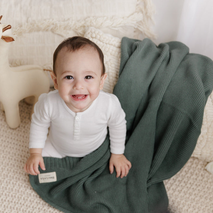 Baby in a white outfit lying on a Hunter green knit blanket, designed by Bleu La La, the Heritage Knit Blanket in 100% Organic Cotton