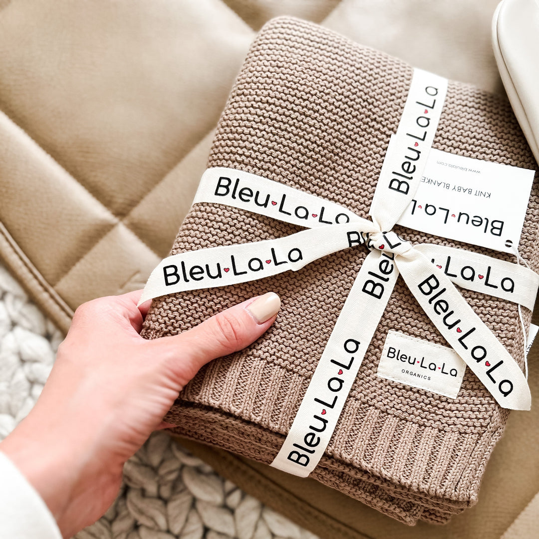 Folded Oatmeal Heritage Knit Blanket, designed by Bleu La La, held by by a hand against a beige couch