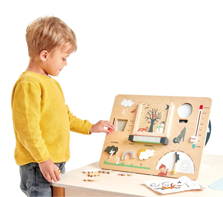 A boy in a yellow sweater reaches to explore the Tender Leaf Toys Weather Watch at the table