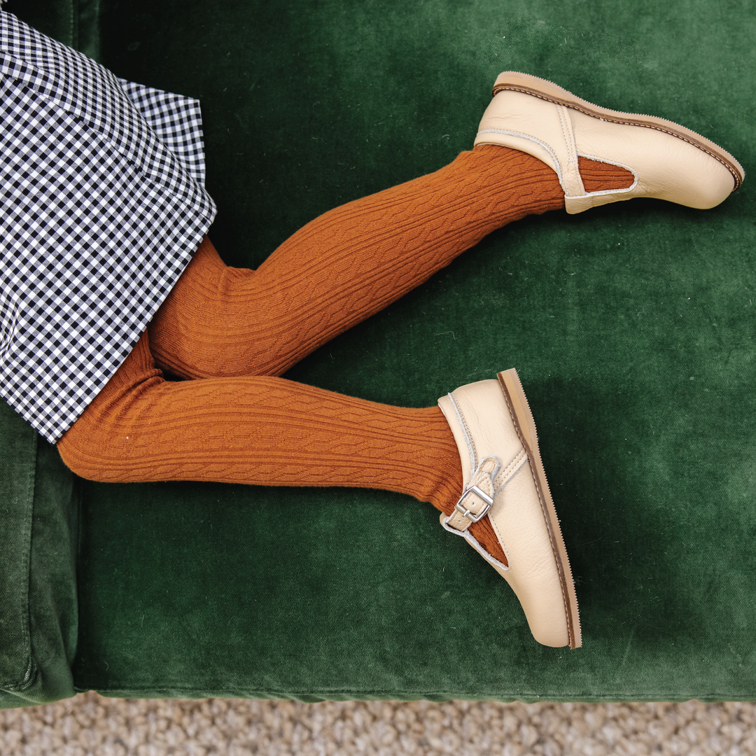 Toddler on a green sofa wearing Little Stocking Co. Sugar Almond Cable Knit Tights with gingham and mary janes