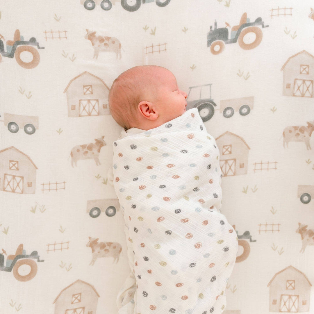 infant wrapped in dot patterned crane baby swaddle on farmhouse tractor barn and cow print baby sheet