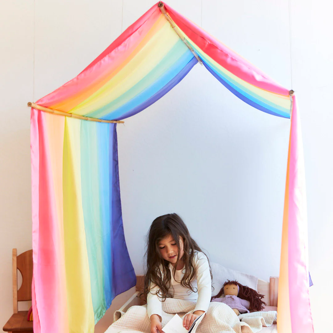 Tucked beneath a rainbow canopy, a child reads, cocooned in the cozy Double Rainbow Playsilk