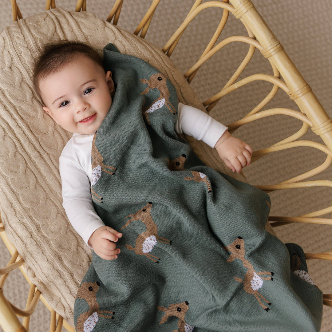 baby in bassinet smiling at camera covered in deer patterned dark green blulala newborn blanket 
