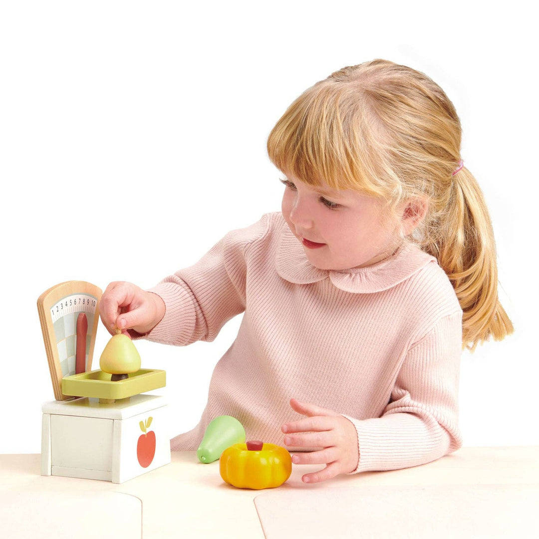 Toddler girl reaches for a piece of wooden fruit beside the Market Scales toy by Tender Leaf Toys