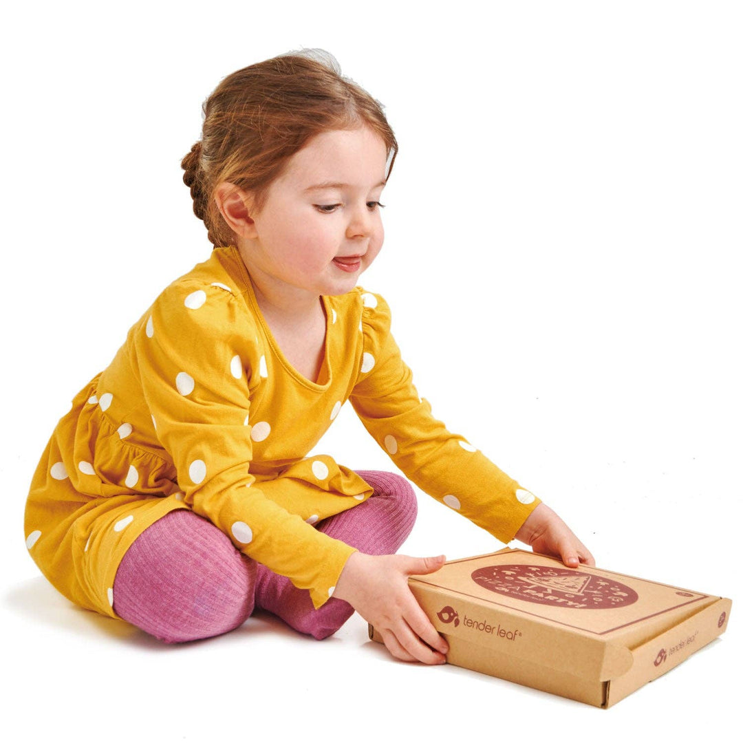 Toddler girl sits cross legged unpacking her new Tender Leaf Toys Pizza Party