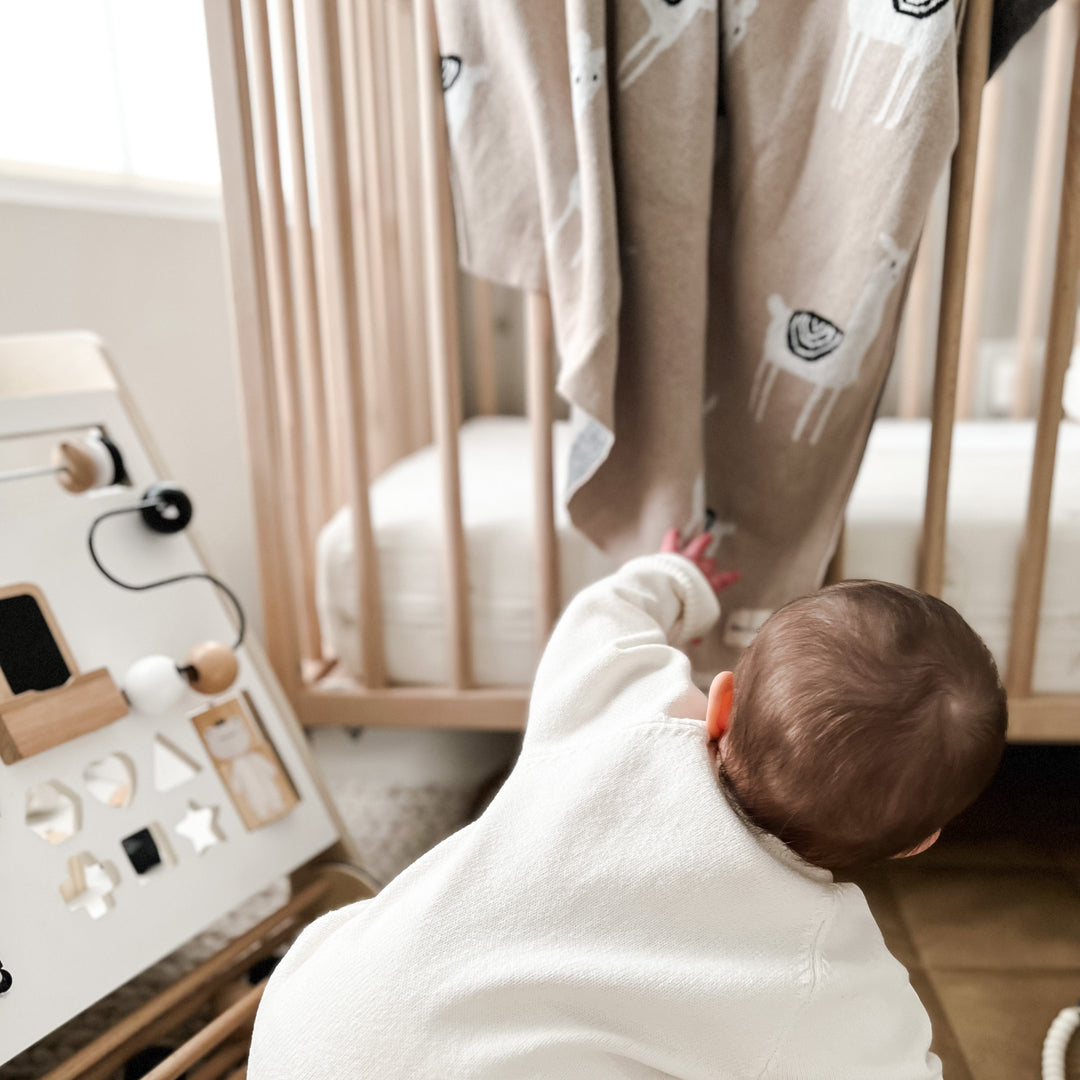 Baby in a white outfit reaching towards a Bleu La La Llama Blanket in Camel