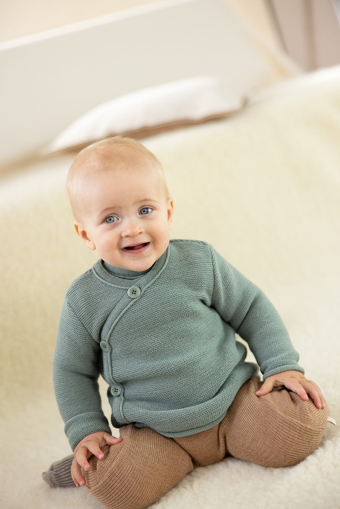 Smiling baby sitting in the Jade Disana Organic Merino Wool Baby Wrap Jacket and tan knit pants, cozy layering for infants