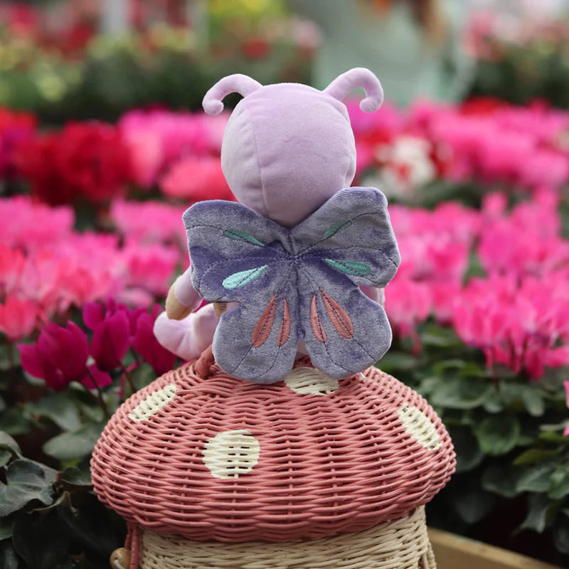 olli ella baby safe stuffed doll sitting on rhattan mushroom basket in buttewrfly outfit looking at flowers 