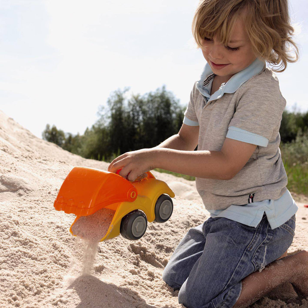 Child playing with an orange-and-yellow wooden excavator toy in the sand.