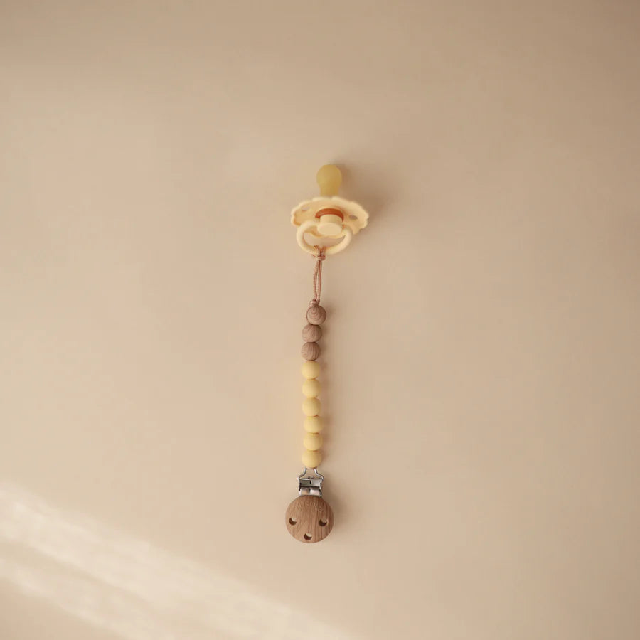 Soft silicone Mushie Eva Pacifier Clip in Mustard Yellow offers a safe and clean pacifier holder.