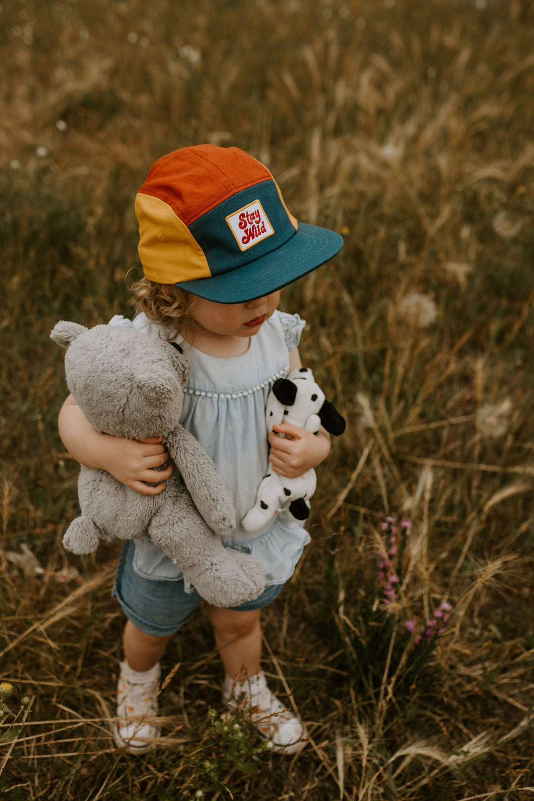 Little girl wears her camp hat while holding a stuffed animal in a sunny grassy field