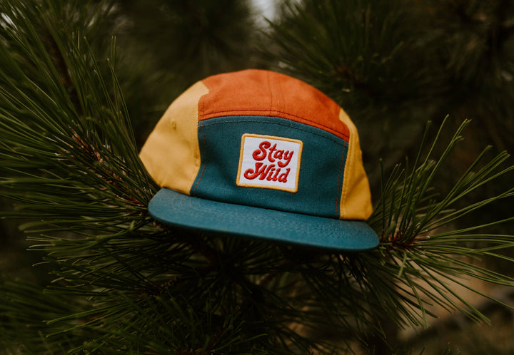 Stay Wild hat hangs from a pine branch outdoors, showing off its cotton five panel design