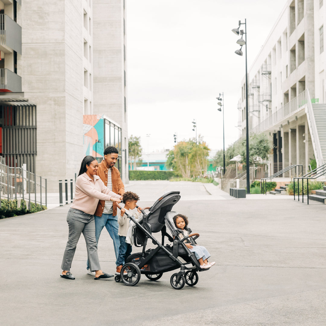 Family with a Uppababy vista v3 travel system stroller and rumbleseat v3 and piggyback rider board walking on a city street.