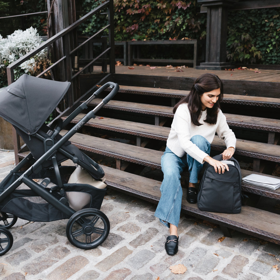 Woman sitting on steps with a black backpack and uppababy vista v3 stroller in jake nearby