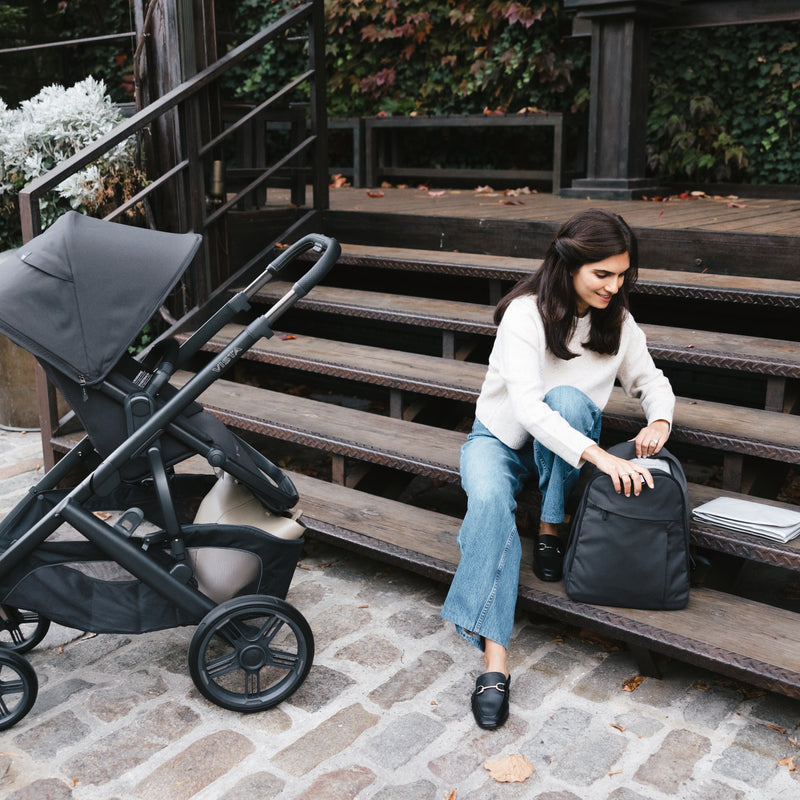 Woman sitting on steps with a black backpack and uppababy vista v3 stroller in jake nearby