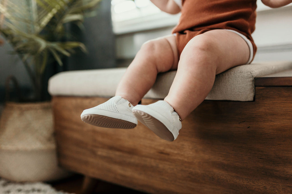 Baby sitting on couch, wearing a pair of Little Love Bug co white quinn shoes