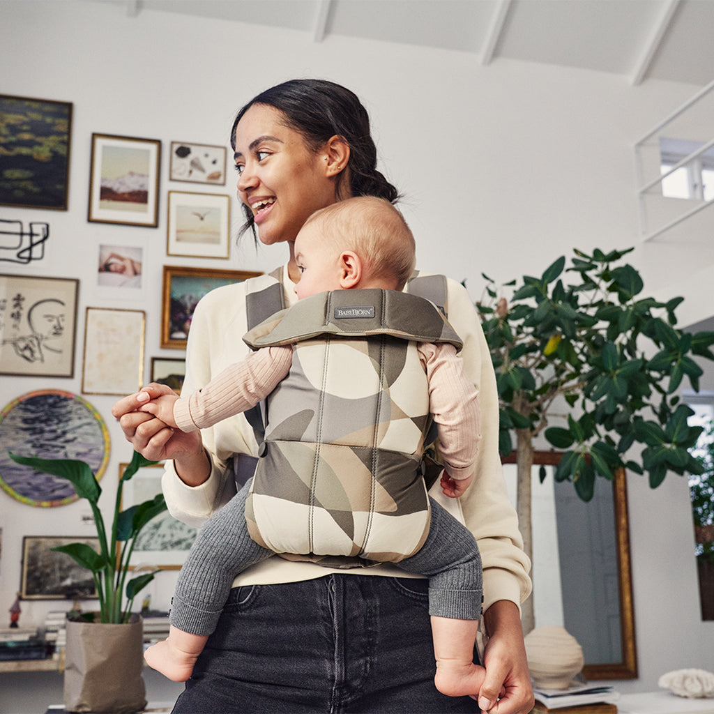 mom smiling in living area with infant in babybjorn khaki green baby carrier