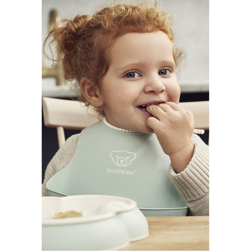 Child Eating and Wearing Babybjorn Soft Feeding Bib Set in Green, with a spill-catching pocket for convenience