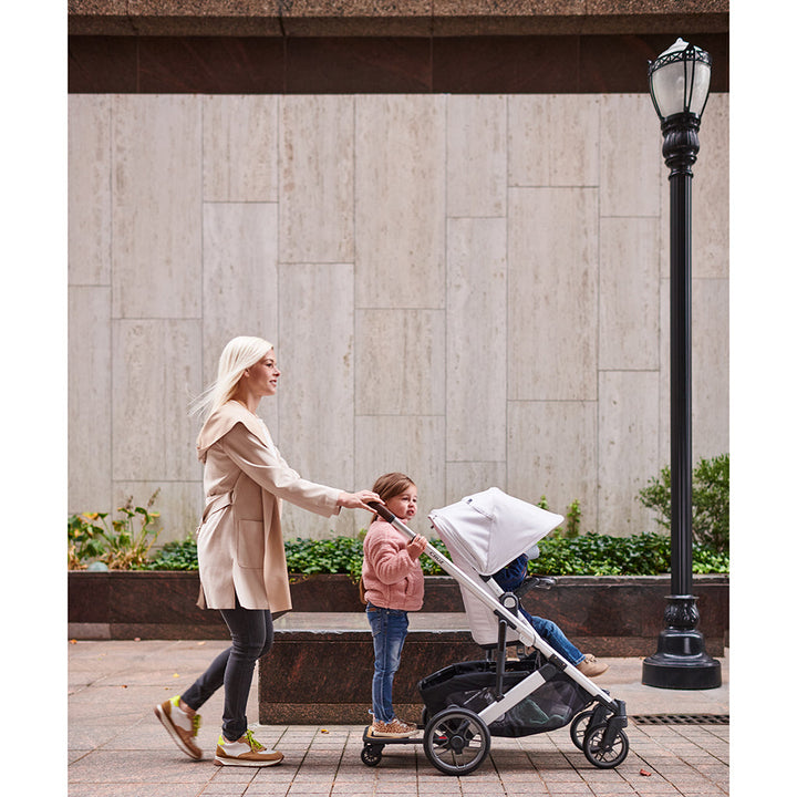 Mom pushing siblings in Bryce Grey Uppababy Cruz V2 Stroller with Sibling Board Accessory