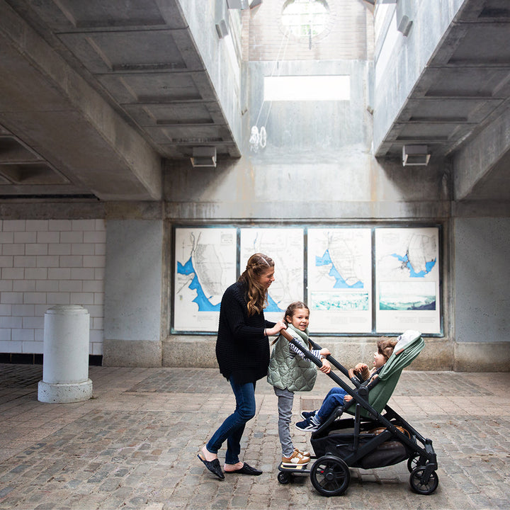 Mom pushing kids in Green Uppababy CRUZ V2 Stroller with attached PiggyBack Sibling Board