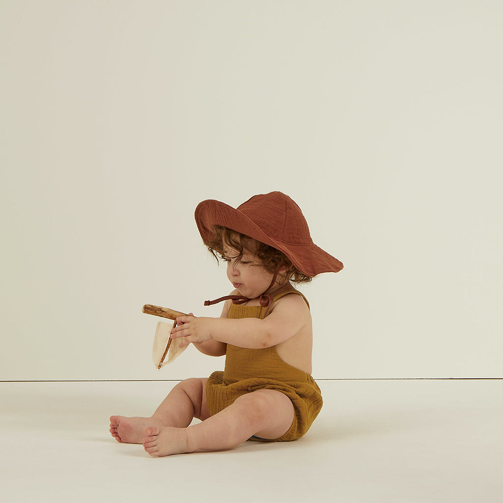 lifestyle_1, criss cross cotton crepe romper is a comfortable one piece perfect for your little one. Child sitting on ground wearing sun hat and romper. 