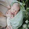 baby wrapped in sage green blanket held by mom next to white flowers