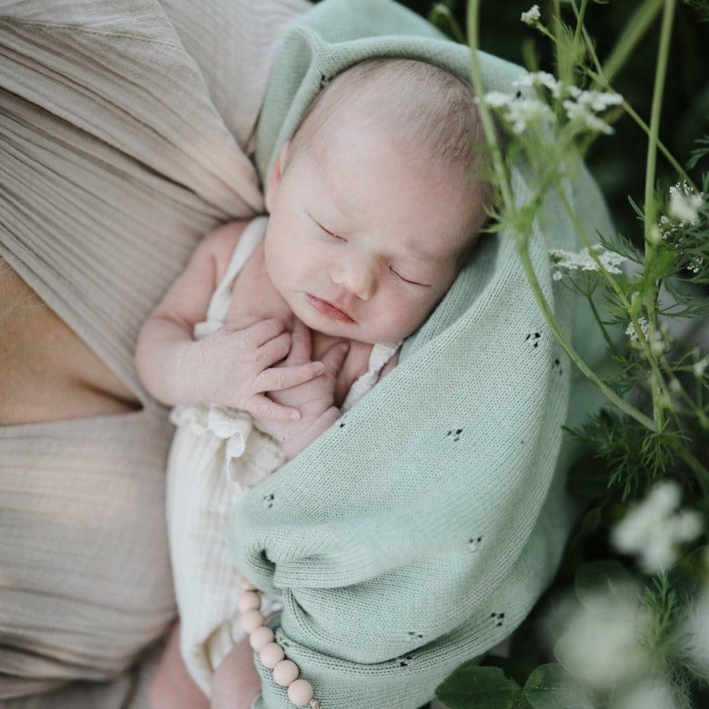 baby wrapped in sage green blanket held by mom next to white flowers