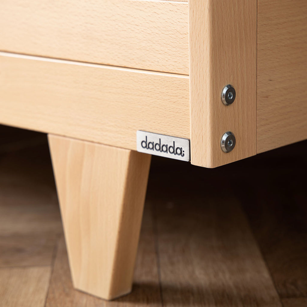 Up close image of dadada logo plate and screws on base of crib. Natural wood crib